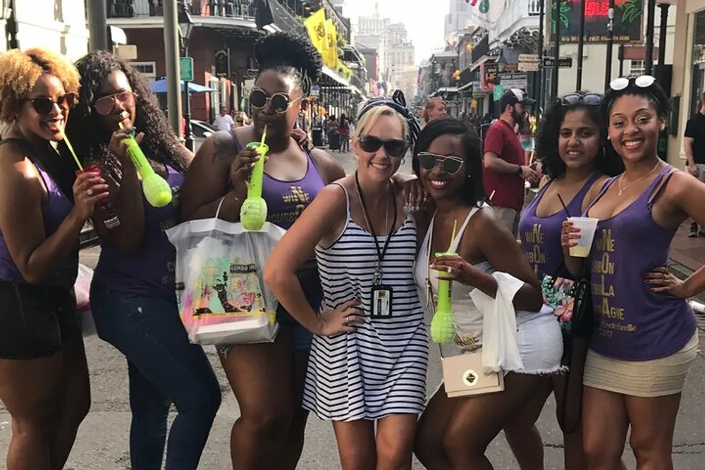 A group of smiling women are enjoying themselves on a sunny street some holding distinctive green drinks and wearing matching purple shirts that suggest they might be participating in a festive event