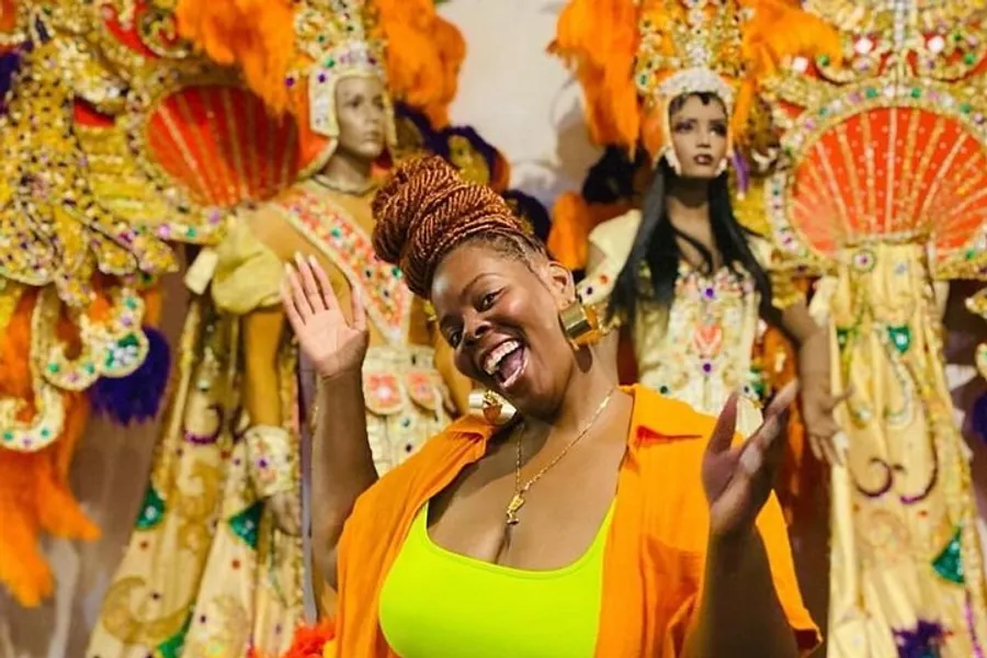 A cheerful person poses in front of a colorful display of ornate carnival costumes.