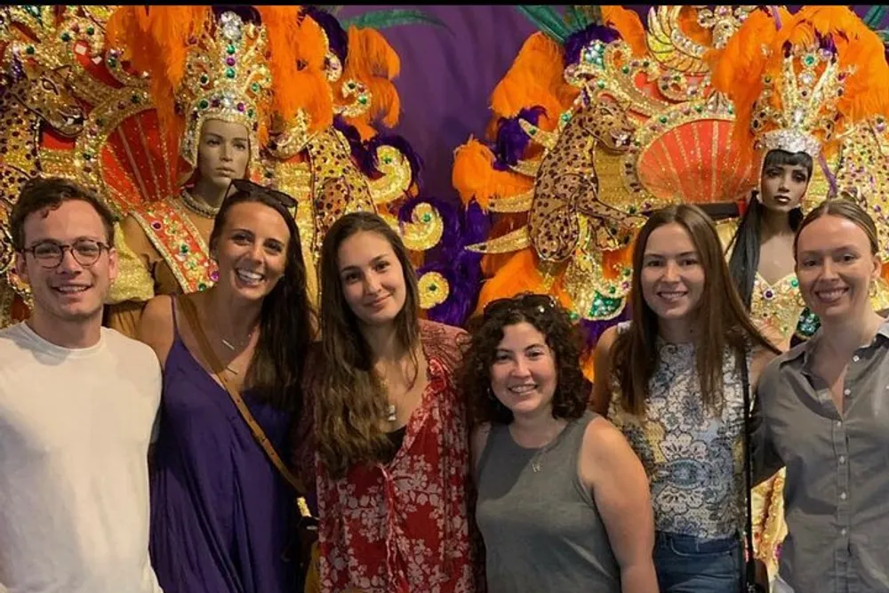 A group of six smiling people are posing for a photo in front of an ornate colorful feathered backdrop reminiscent of a carnival or festival