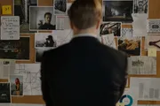 A person is seen from behind, contemplating a cork board filled with various photographs, notes, and strings that suggest a complex investigation or conspiracy.