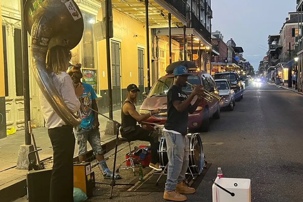 A trio of street musicians performs on a lively city street in the evening with one playing a tuba another on drums and a trumpet player all contributing to what appears to be a vibrant music scene