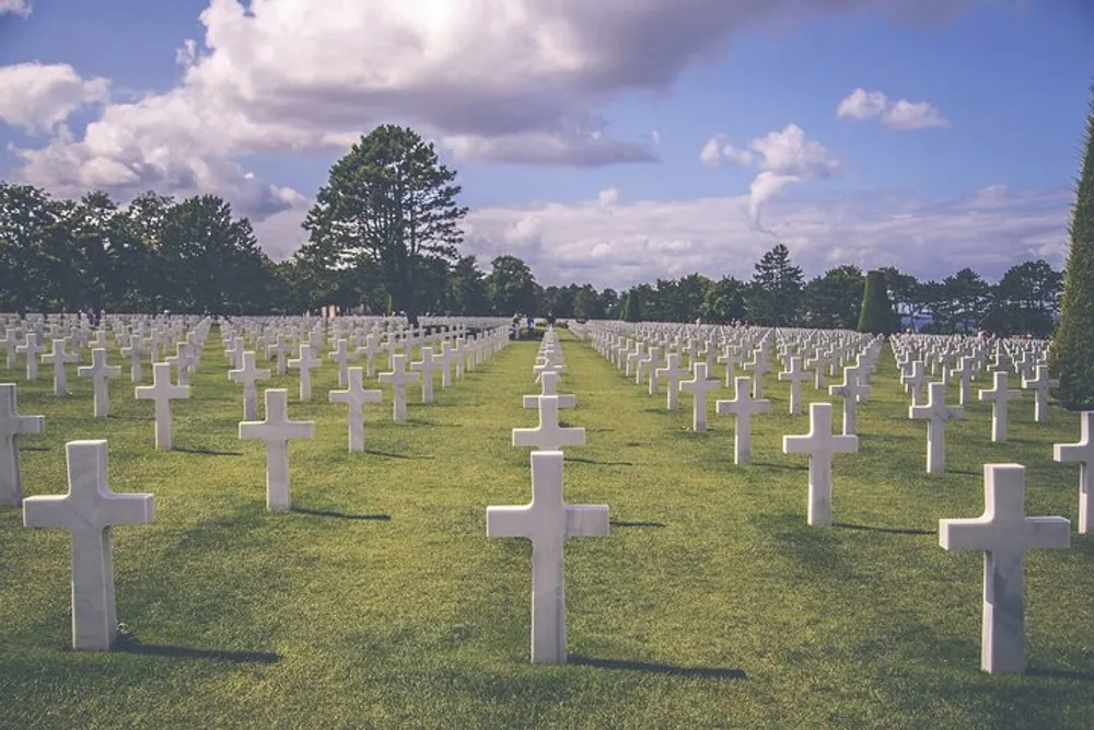 The image shows a serene and orderly military cemetery with rows of white crosses marking the graves against a backdrop of trees and a cloudy sky