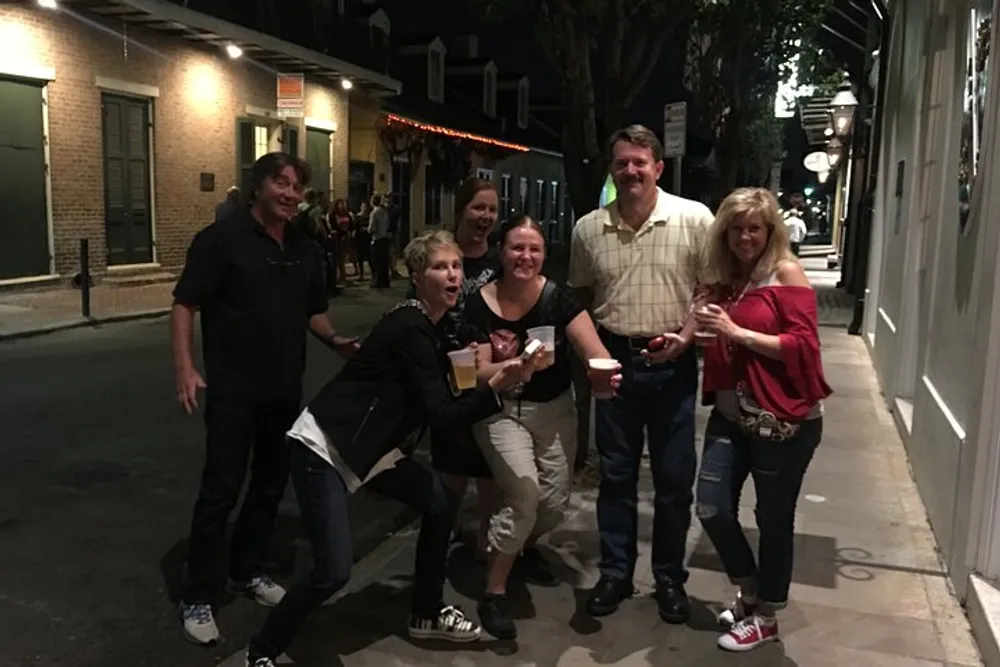 A group of people appear to be enjoying themselves at night on a city street with some holding beverages and demonstrating playful poses for the photo