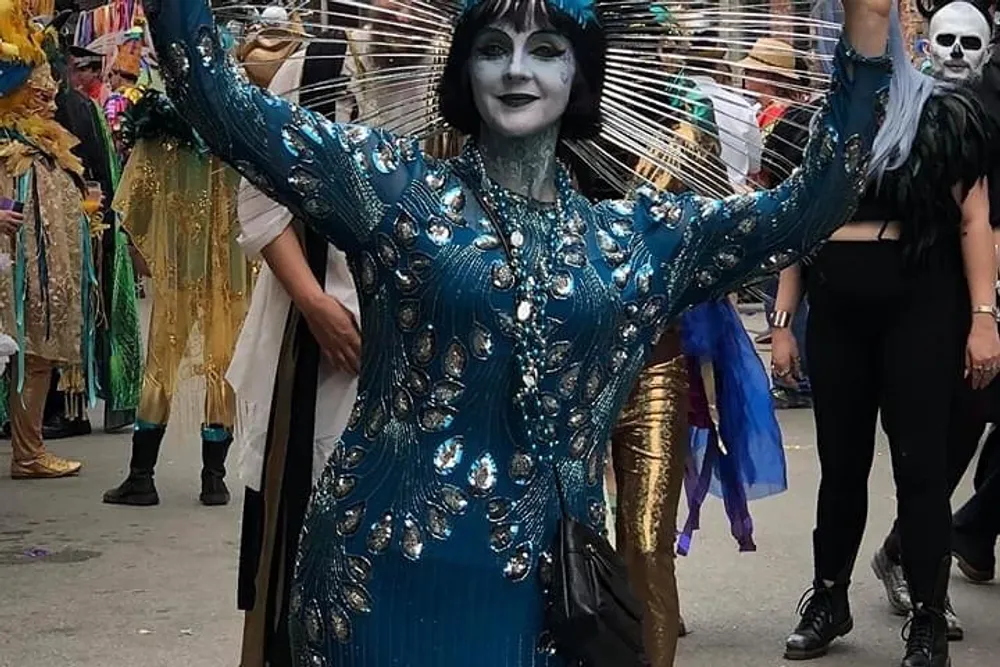 A person in an elaborate blue and silver costume with face paint is posing with outstretched arms amidst what appears to be a festive parade or event