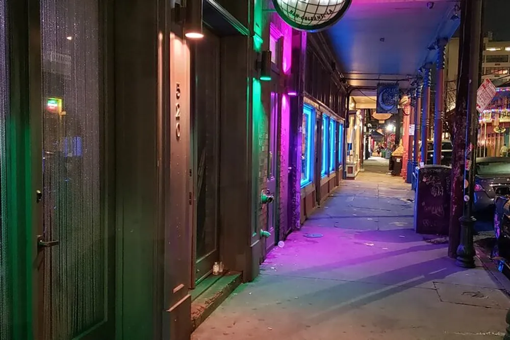 The image depicts a narrow urban alley at night illuminated by vibrant neon lights casting purple and blue hues on the pavement and buildings