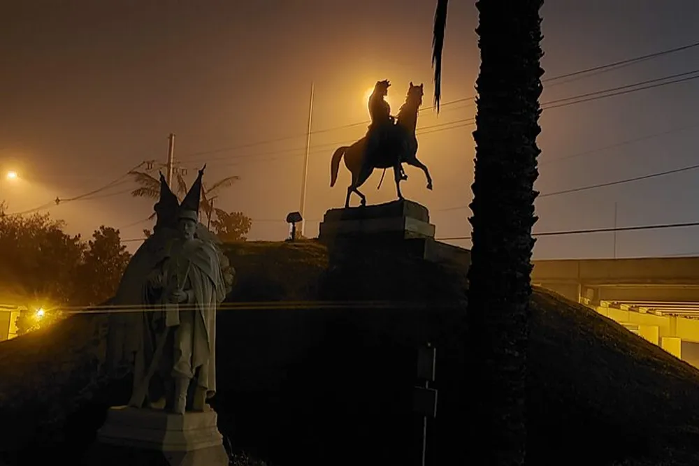 A silhouette of an equestrian statue is set against a night sky illuminated by artificial lights with another statue visible in the foreground