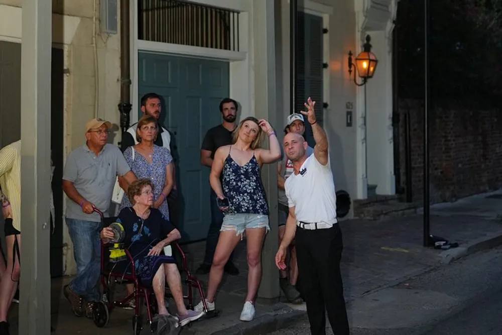 A group of people including a man who appears to be a tour guide gesturing upwards are gathered on a street possibly listening to an explanation or participating in a walking tour