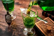 A green liquid, likely absinthe, is being poured into a glass over a slotted spoon with a sugar cube, a traditional preparation method for this potent spirit.