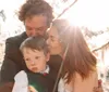 A child is lovingly embraced by two adults in an outdoor setting with sunlight streaming through the trees
