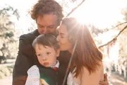 A child is lovingly embraced by two adults in an outdoor setting with sunlight streaming through the trees.