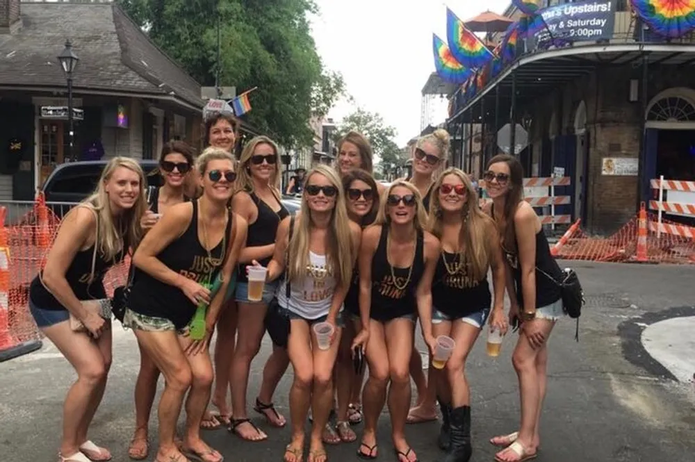 A group of happy women are posing together on a city street several of them wearing matching tank tops suggesting a group celebration or event