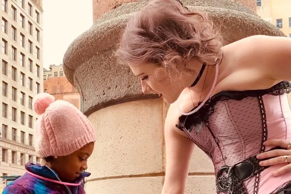 A woman in a pink corset is bending down to look at a young child wearing a pink beanie and a blue jacket with both standing on a city street