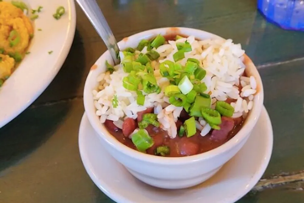 A bowl of red beans and rice topped with chopped green onions a classic Southern dish is presented on a table with a fork on the side and a plate with another dish partially visible in the background