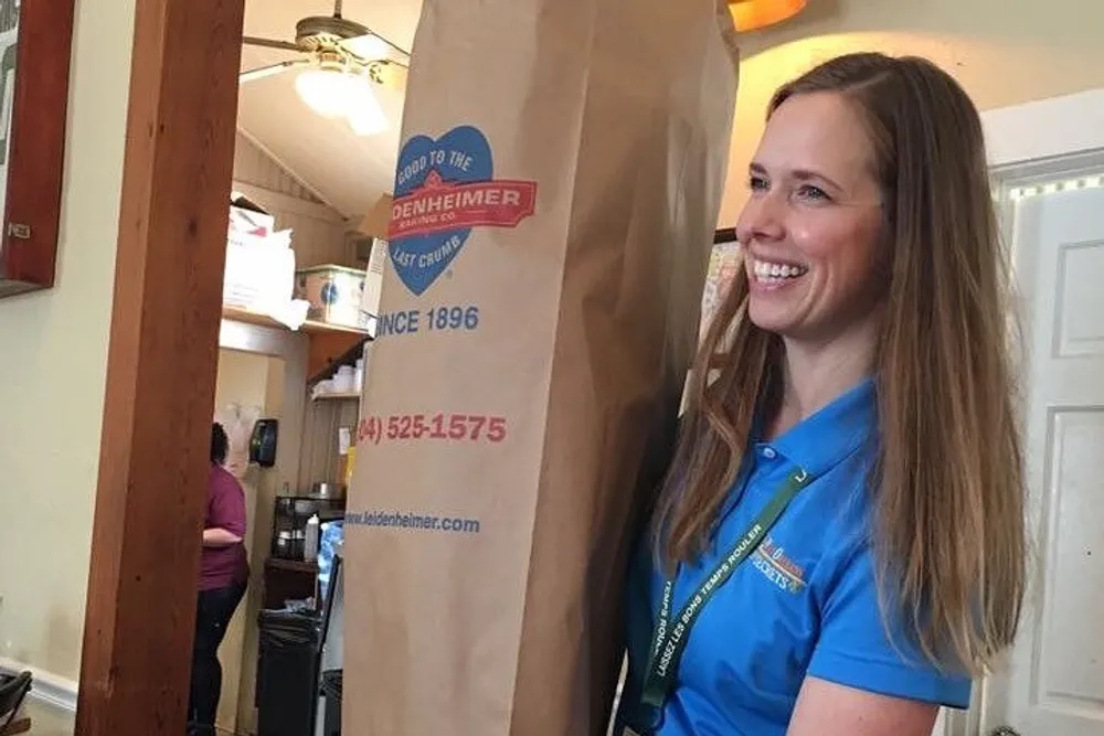 A smiling woman in a blue shirt is standing next to a large paper bag with a logo and text on it in a room that appears to be a commercial setting