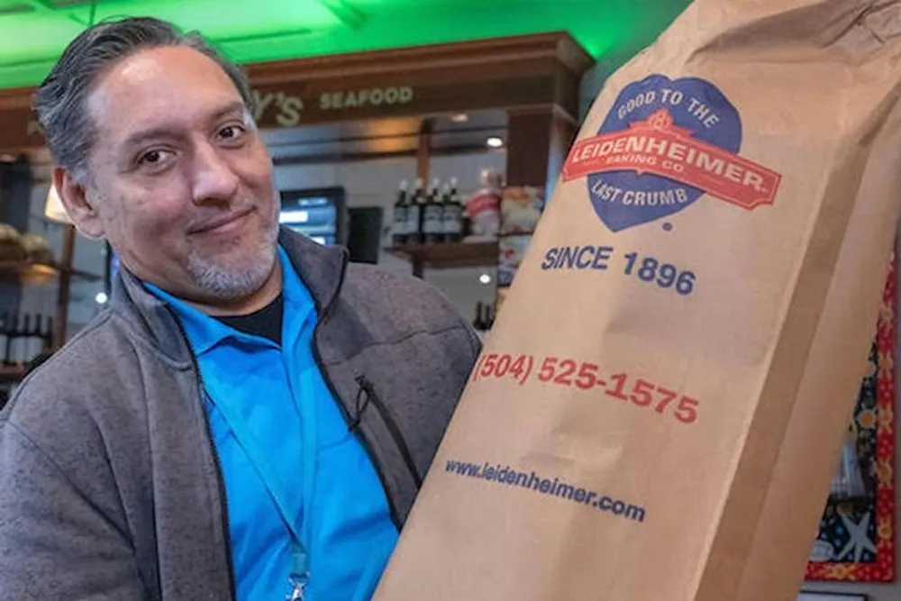 A man is smiling while holding a large paper bag from the Leidenheimer Baking Company that reads Good to the Last Crumb and displays a since 1896 date