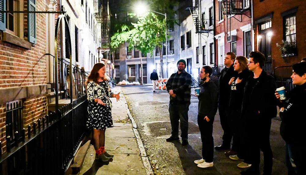 A group of people is attentively listening to a woman speaking on a dimly lit urban street at night