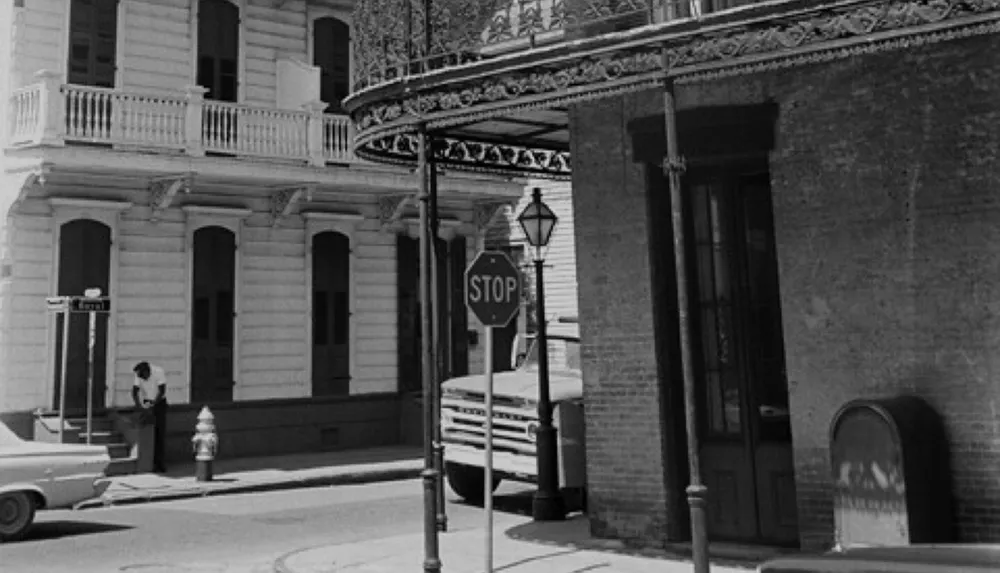 The image is a black and white street scene showing a person walking past a building with wrought-iron balconies next to a stop sign and a parked vehicle evoking a sense of historical ambiance