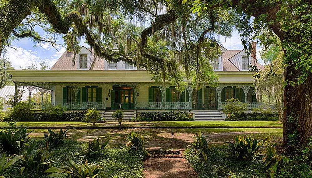 This image shows a picturesque two-story plantation-style house with a large front porch flanked by lush greenery and hanging Spanish moss