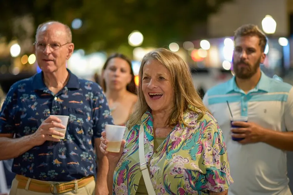 A group of adults each holding a drink appear to be engaged in a lively outdoor event with a focus on a smiling woman in a colorful blouse