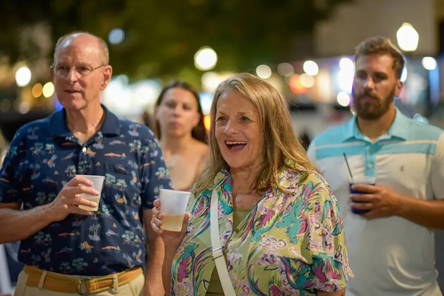 A group of adults, each holding a drink, appear to be engaged in a lively outdoor event, with a focus on a smiling woman in a colorful blouse.