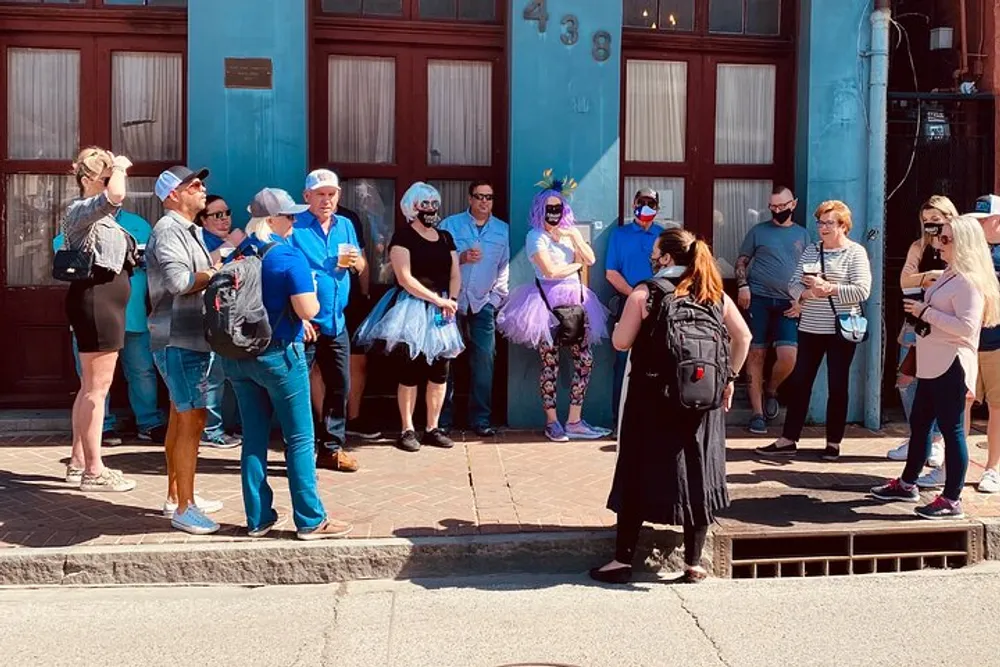 A diverse group of people some in casual attire and others wearing tutus and fanciful accessories are gathered on a sidewalk engaging in social interactions on a sunny day