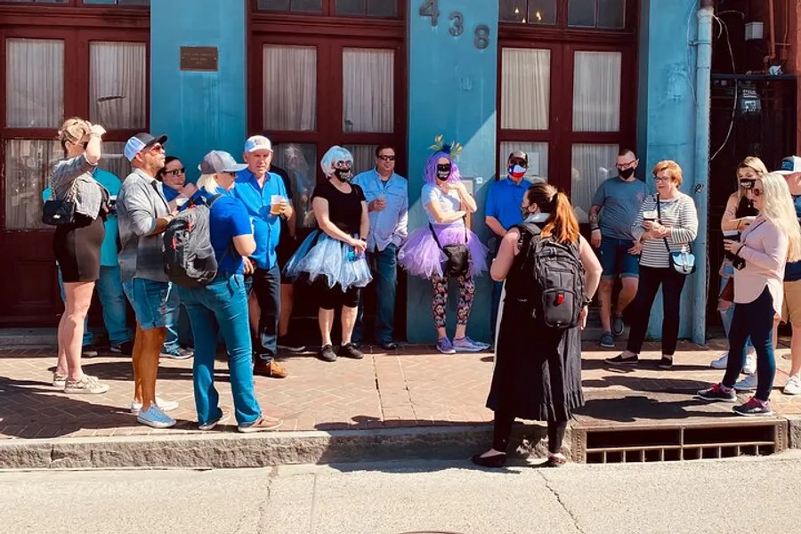 A diverse group of people, some in casual attire and others wearing tutus and fanciful accessories, are gathered on a sidewalk, engaging in social interactions on a sunny day.