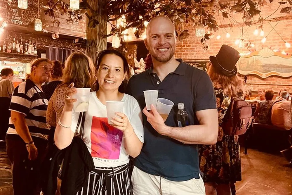 Two people are smiling and holding plastic cups seemingly enjoying a social event at a venue decorated with eclectic wall art and a festive atmosphere