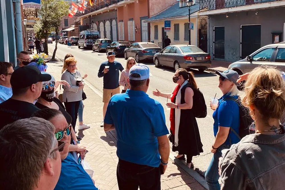 A group of people is gathered on a sunny street attentively listening to a woman who appears to be guiding a walking tour