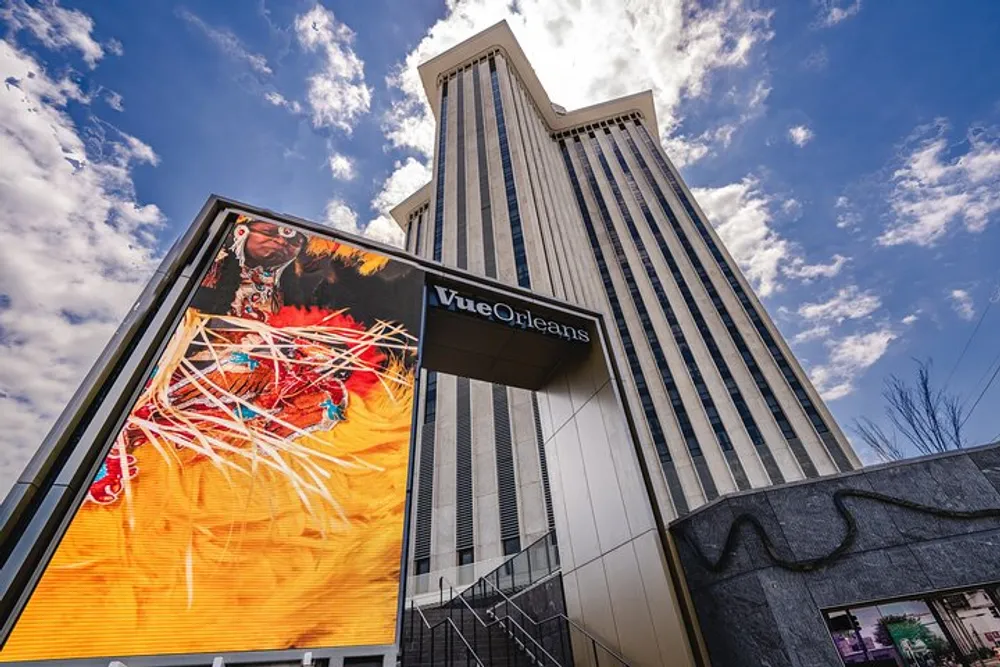 The image shows a tall building with a large digital display screen featuring a vibrant graphic set against a backdrop of a partly cloudy sky