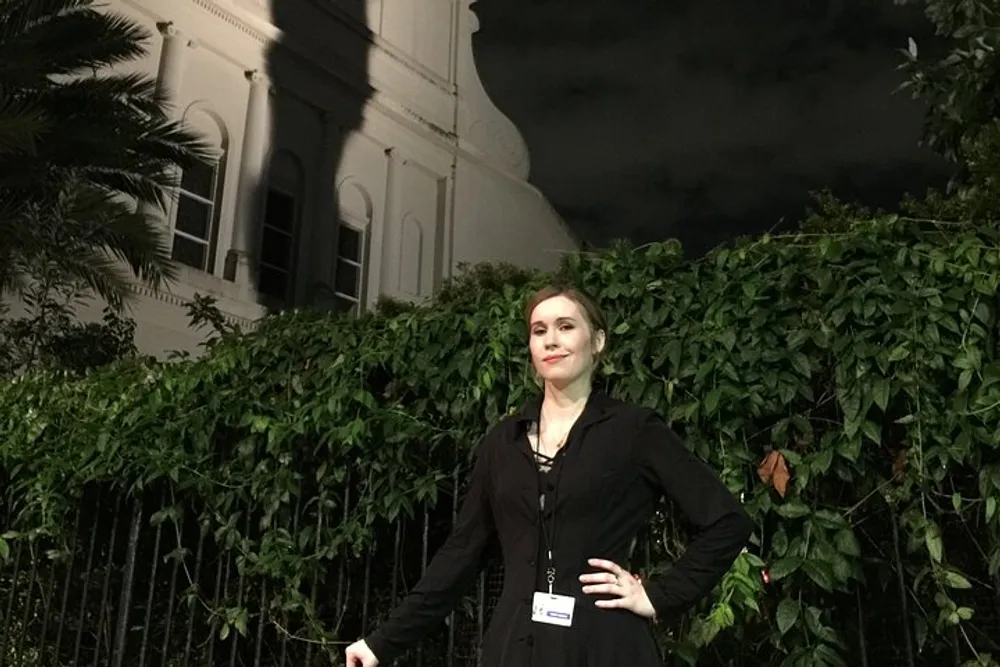 A person is standing confidently at night in front of a vine-covered fence with a historic-style building and dark sky in the background