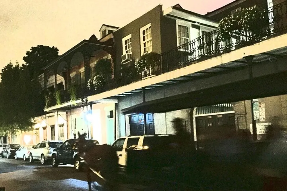 This is an image of a dimly lit street at night with parked cars and a building featuring balconies with intricate railings likely taken with a high ISO or under low light conditions resulting in noticeable graininess in the photo