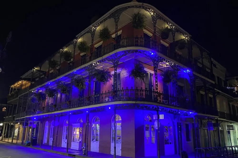 A historic building illuminated with blue lighting at night featuring ornate wrought-iron balconies and lush greenery