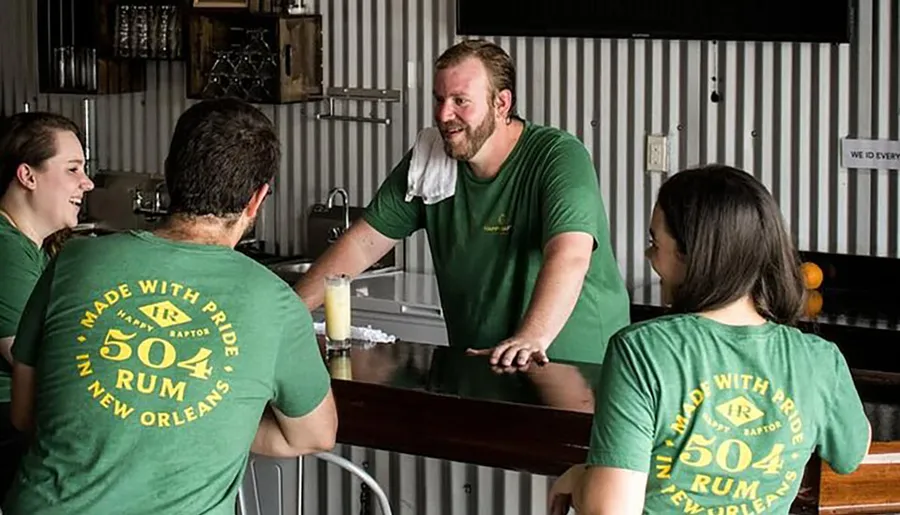 A group of people, wearing matching green shirts advertising '504 Rum New Orleans,' are chatting and smiling around a bar area with a metal backdrop.