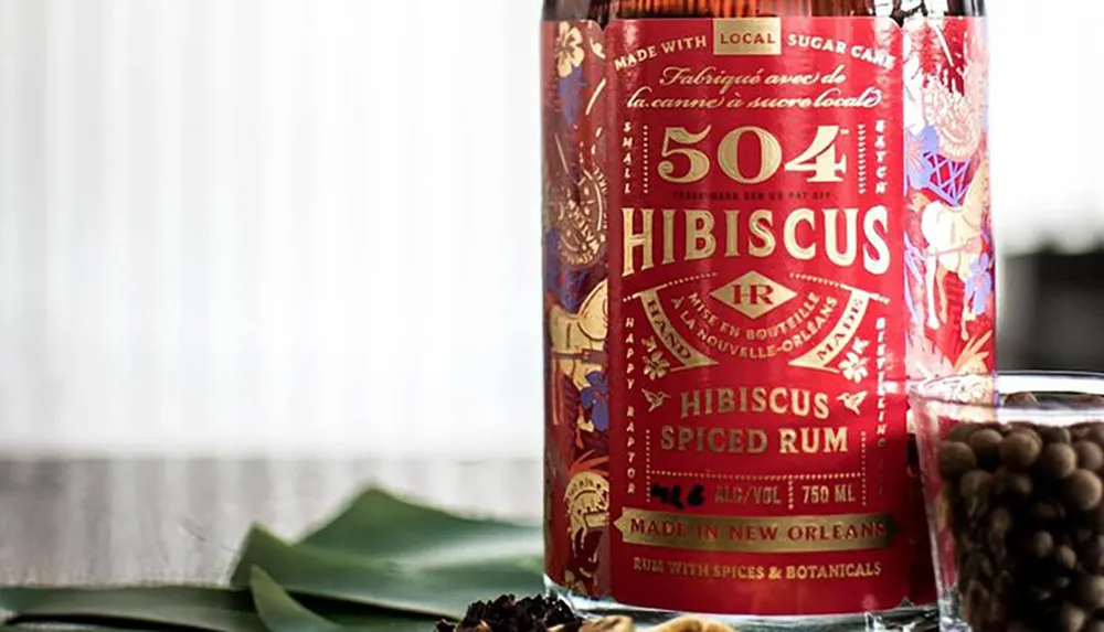 The image features a bottle of 504 Hibiscus Spiced Rum with intricate labeling positioned next to green leaves and in front of a glass containing what appears to be spices or botanicals