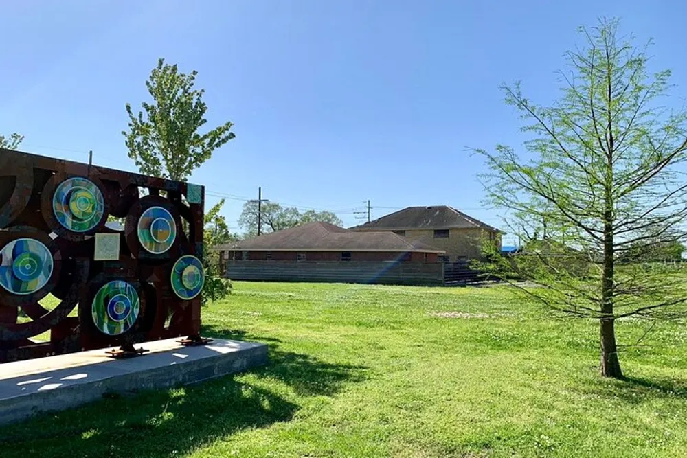 A sunny day showcases an outdoor setting with an artistic structure featuring circular targets beside a young tree with a house and clear blue sky in the background