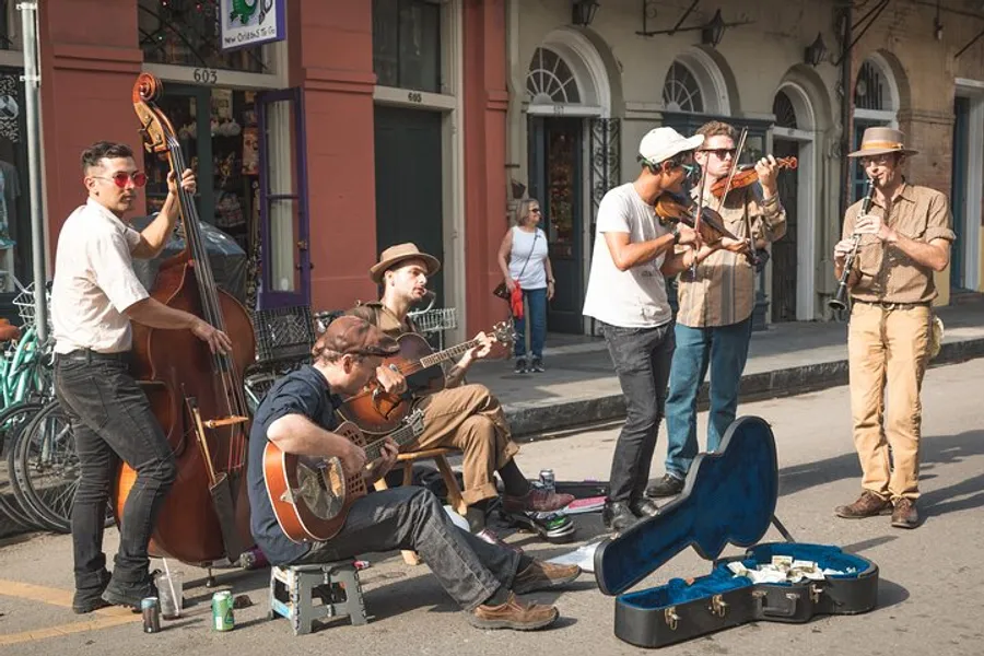 A group of street musicians is performing live on a city sidewalk, playing an upright bass, acoustic guitars, and a violin, with an open guitar case in front for tips.