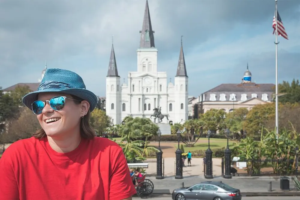 A smiling person wearing a blue striped hat and sunglasses is pictured in the foreground with the iconic Saint Louis Cathedral and Jackson Square in the background under a cloudy sky