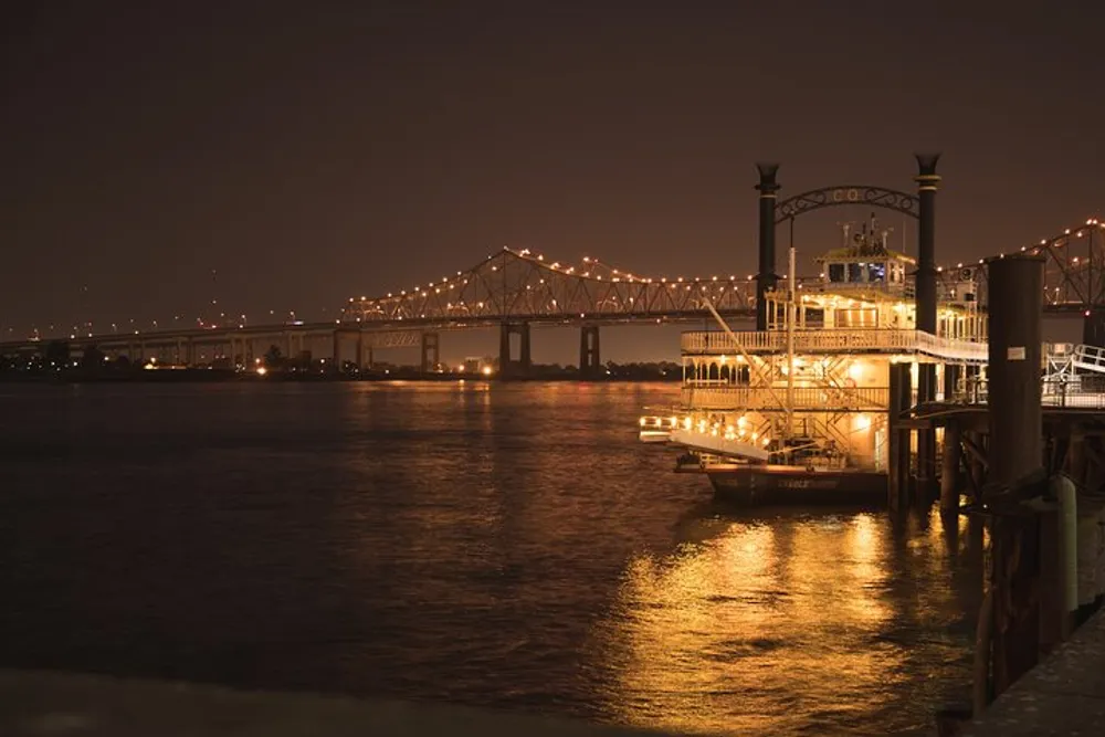 A paddle steamer is docked at a pier at night with a lit bridge in the background