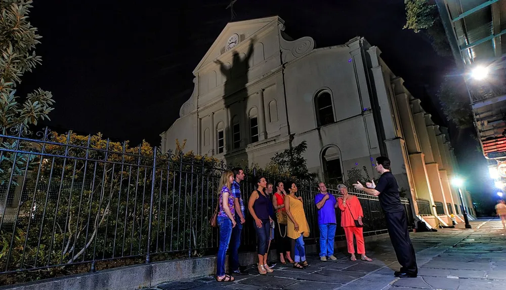 A group of people listens to a guide during a nighttime tour in front of a historical building with striking architecture