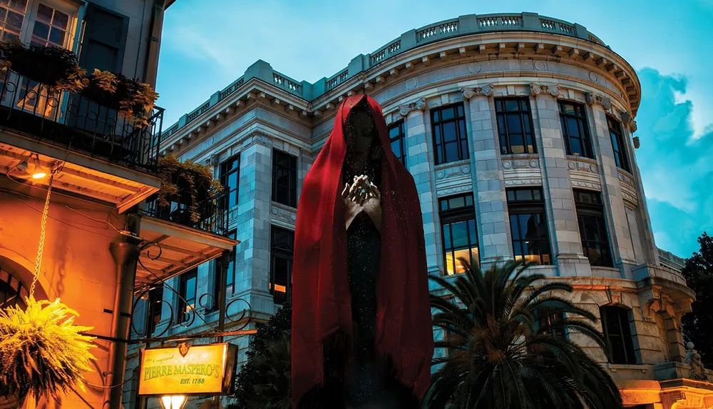 A statue of a robed figure with a red cloak stands prominently before a classically designed building with a balcony and palm trees under an evening sky