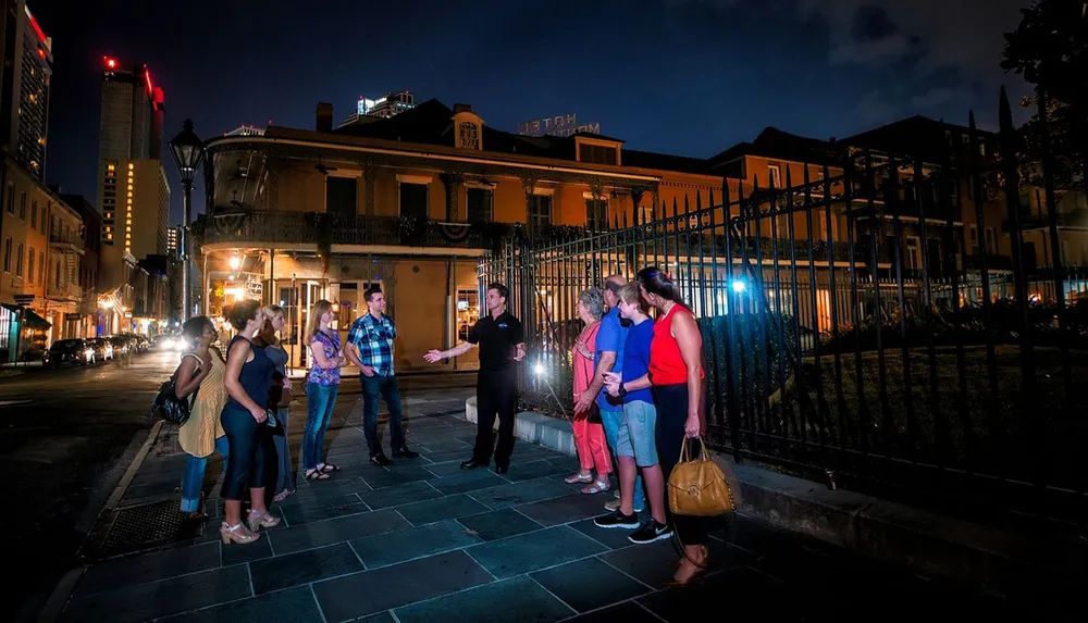 A group of people is engaged in what appears to be a night-time walking tour on an urban street with an individual gesturing and speaking to the attentive group under city lights