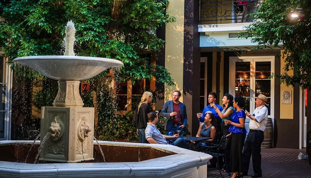 A group of people are socializing around a classic fountain in an open-air courtyard with lush greenery in the background