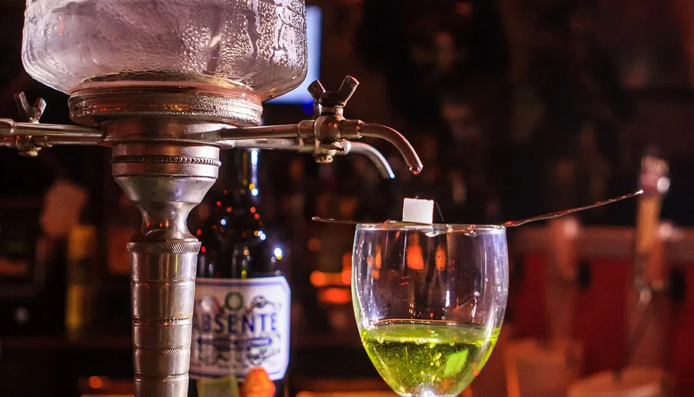 An absinthe fountain is dripping water onto a sugar cube on a slotted spoon over a glass of green absinthe in a warmly lit bar setting