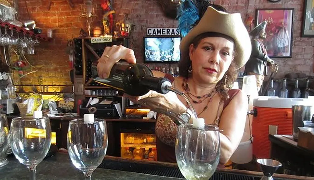 A person wearing a cowboy hat is pouring a bottle of wine into a glass at a bar adorned with various items and a sign that says CASH ONLY