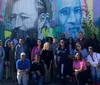 A diverse group of people are posing for a photo in front of a colorful mural featuring portraits