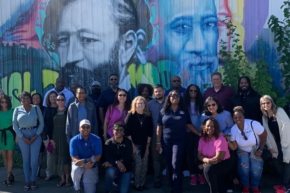 A diverse group of people are posing for a photo in front of a colorful mural featuring portraits