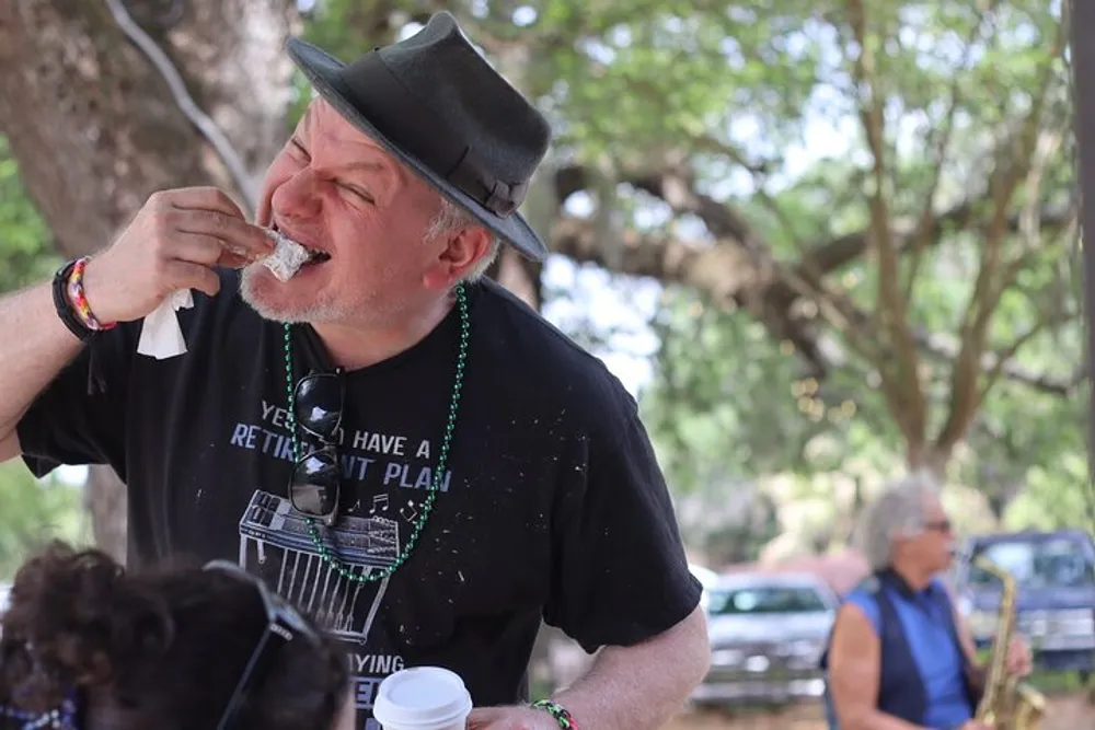 A man wearing a black hat and t-shirt is enjoyably biting into a beignet at an outdoor event where a person can be partially seen playing a saxophone in the background