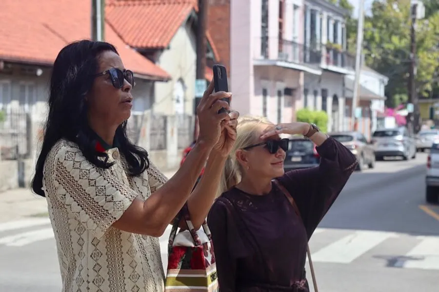 Two women are standing on a street, one taking a picture with her smartphone while the other shields her eyes from the sun.