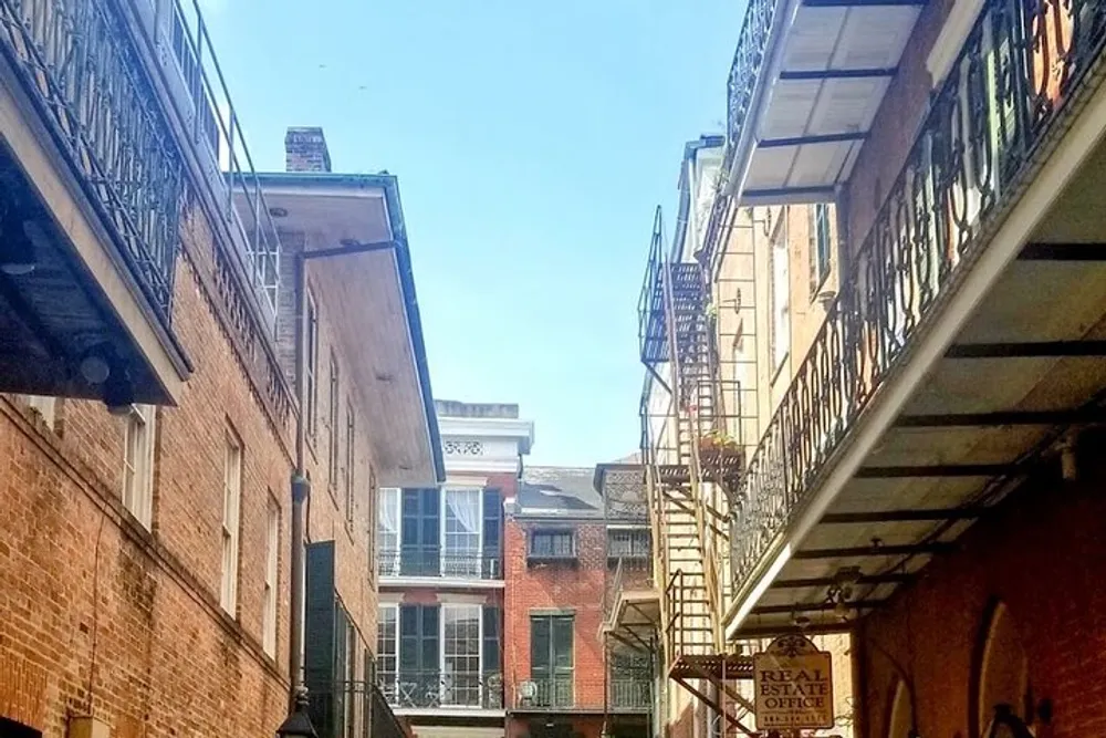 The image shows a sunny view of a charming narrow alley with wrought-iron balconies and fire escapes between old brick buildings suggesting a historic urban environment