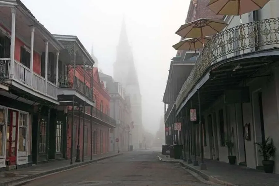 An early morning fog shrouds the historic architecture of what appears to be a street in the French Quarter of New Orleans, with the outline of a church spire barely visible in the background.